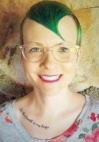 Head shot photo of director Abra Singleton, with shock of vivid florescent green hair and tattoo under collar bone reading, "It's His breath in my lungs" 